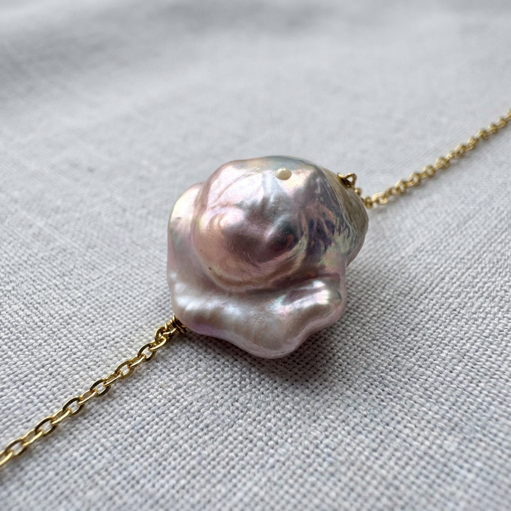 Close-up of a silvery-blush keshi pearl on a gold-over-sterling-silver chain, showing its irregular square outline, raised nacre folds and rose-mauve reflections on grey linen.