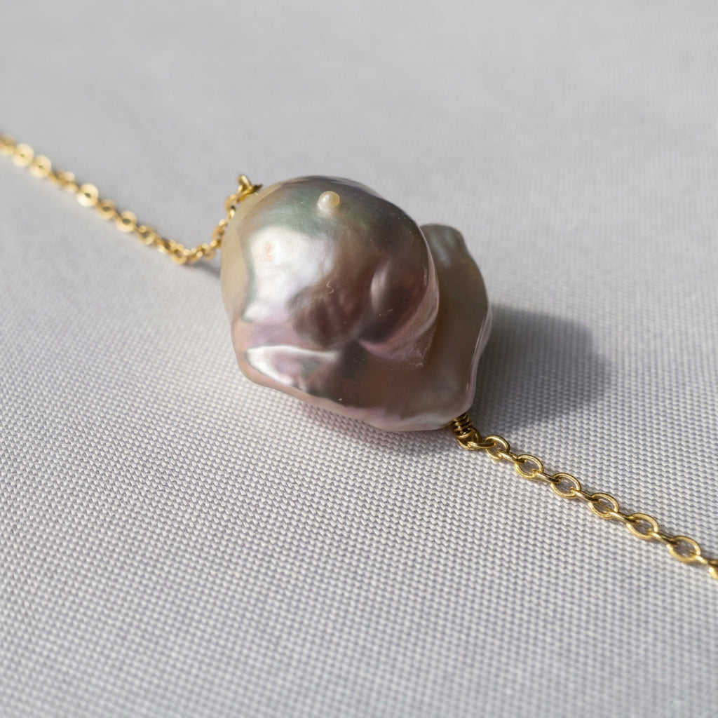 Close-up of a silvery-blush keshi pearl on a gold-over-sterling-silver chain in sunlight, showing its raised rounded centre, folded edges and pink-green iridescence on pale fabric.