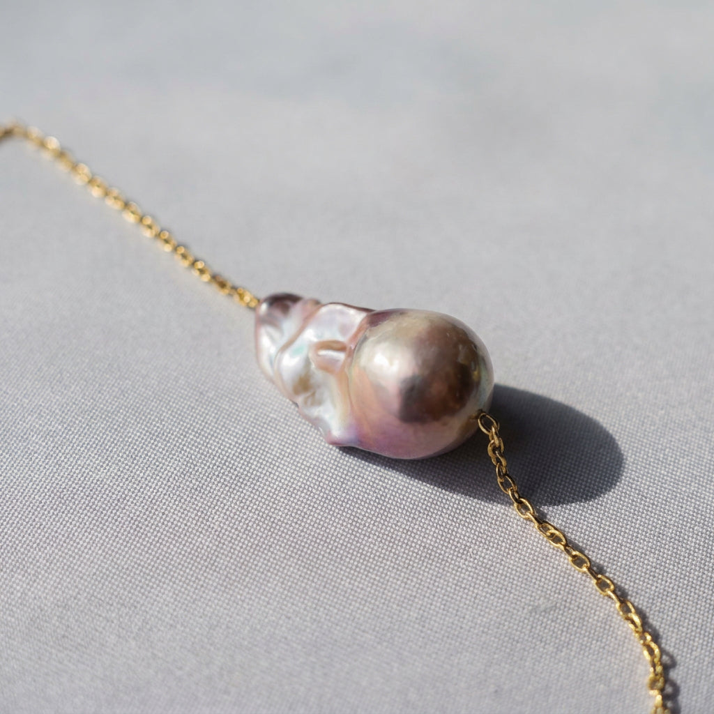 Close-up of a rainbow keshi pearl threaded onto a fine gold-filled chain on grey fabric in sunlight, showing its rounded champagne-pink dome and rippled silver-white nacre folds.