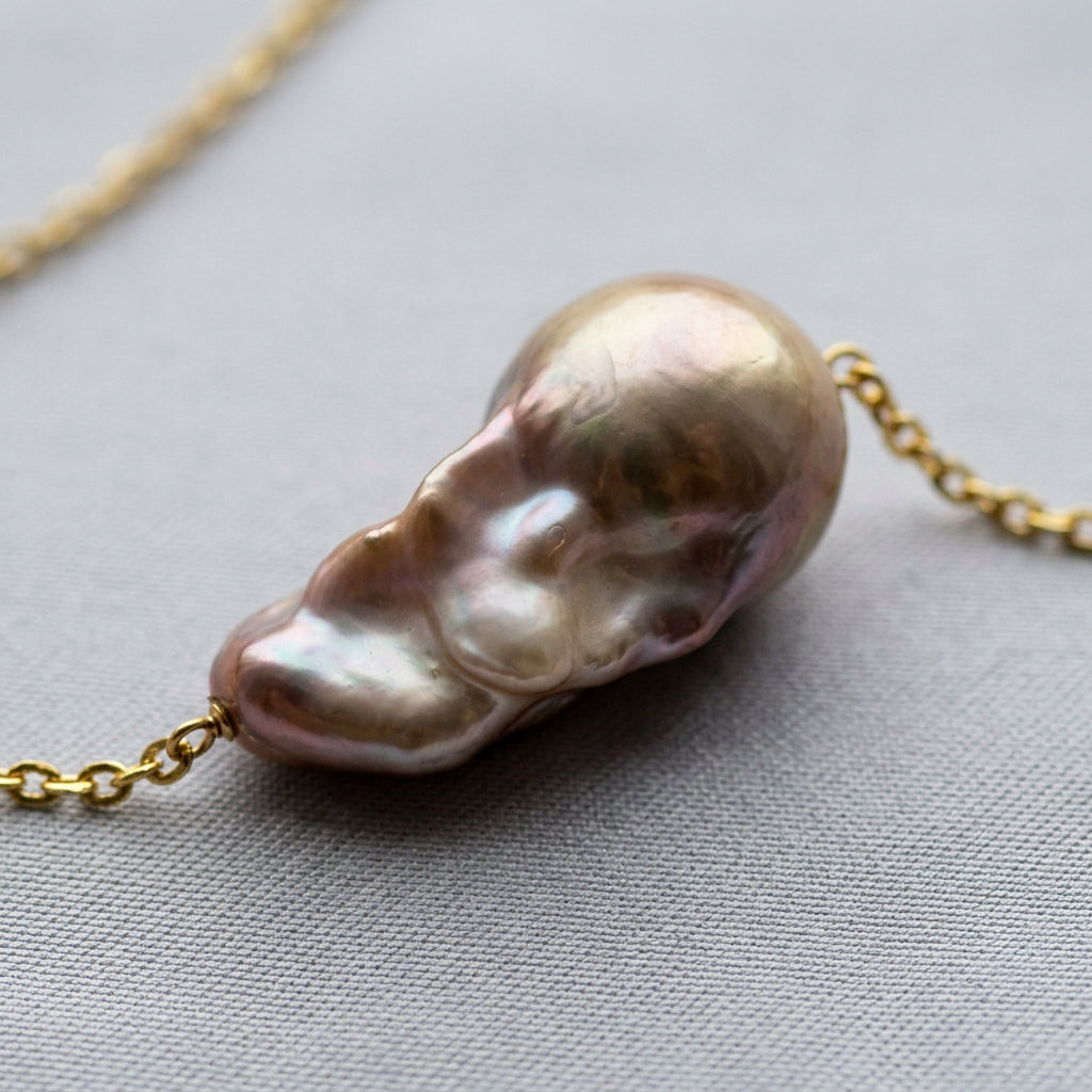 Close-up of a rainbow keshi pearl on a gold-filled chain, showing its rounded champagne dome, sculptural nacre folds and rose, lavender and silver reflections on grey fabric.