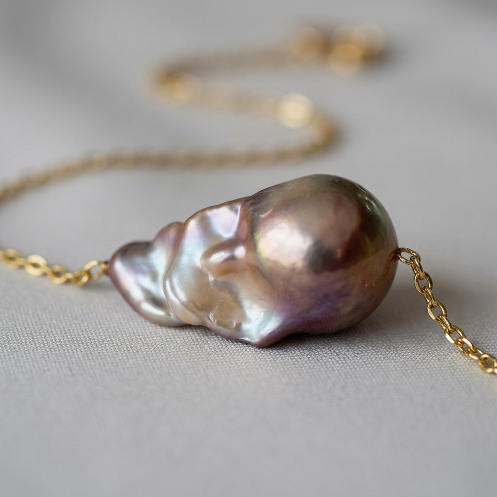 Low-angle close-up of a rainbow keshi pearl on a fine gold-filled chain, showing its bulbous champagne-pink dome and rippled silver-lavender nacre on grey fabric.