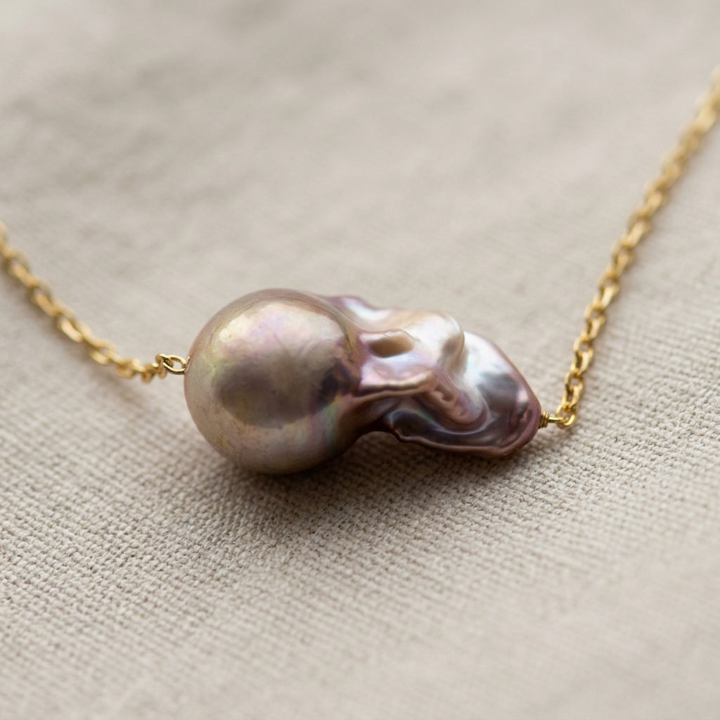 Close-up of a rainbow keshi pearl on a fine gold-filled chain, with its rounded champagne dome to the left and folded rose-lavender nacre to the right on cream linen.