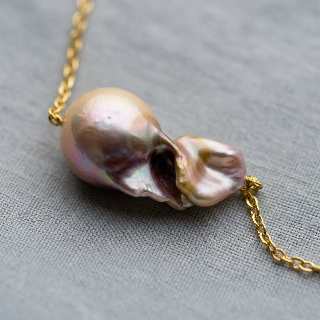Close-up of a rainbow keshi pearl on a gold-filled chain, showing deep sculptural folds and champagne, pink, lavender and silver iridescence on grey linen.