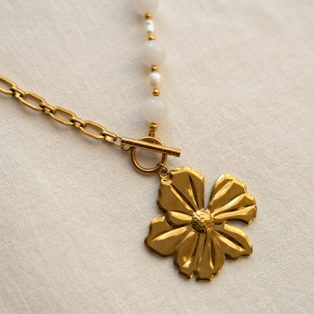 Gold-plated five-petal flower pendant beneath a fastened circular toggle clasp, with paperclip chain on one side and pink quartz beads and white baroque pearls on the other, resting on cream linen.
