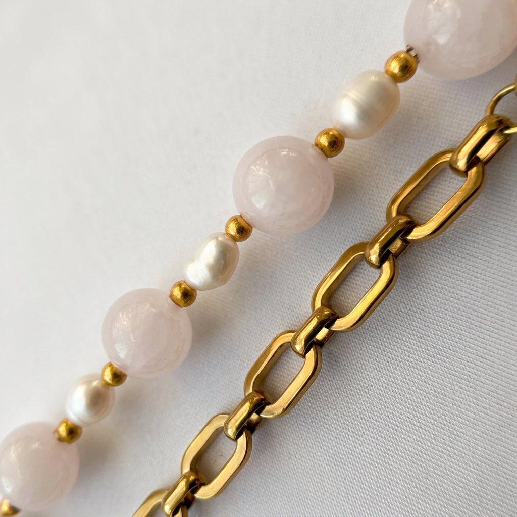 Close-up of faceted pale pink quartz beads alternating with white rice-shaped baroque pearls and gold-plated spacers, beside a gold-plated paperclip chain on white fabric.
