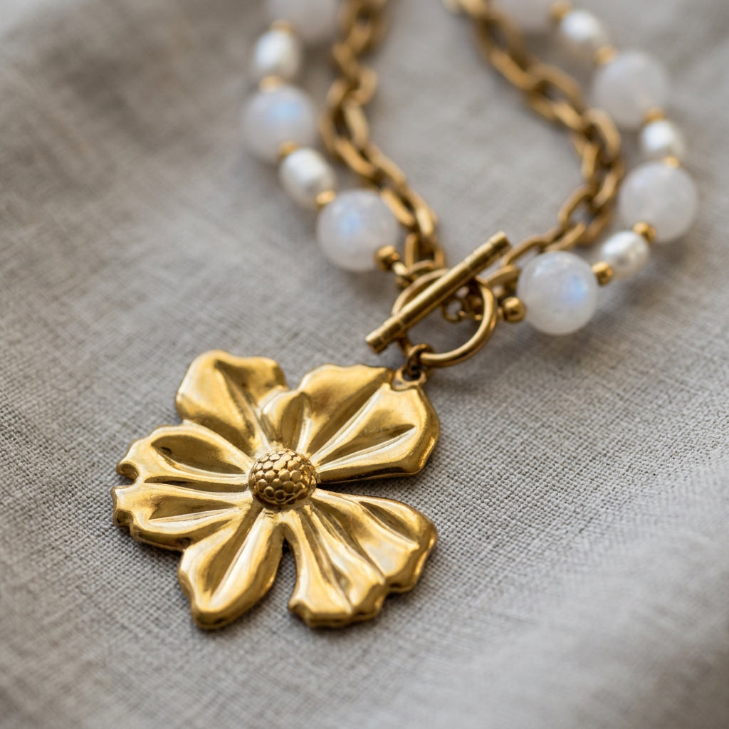 Close-up of a gold-plated five-petal flower pendant with a textured centre and fastened toggle clasp, with pink quartz beads, white baroque pearls and paperclip chain on grey linen.