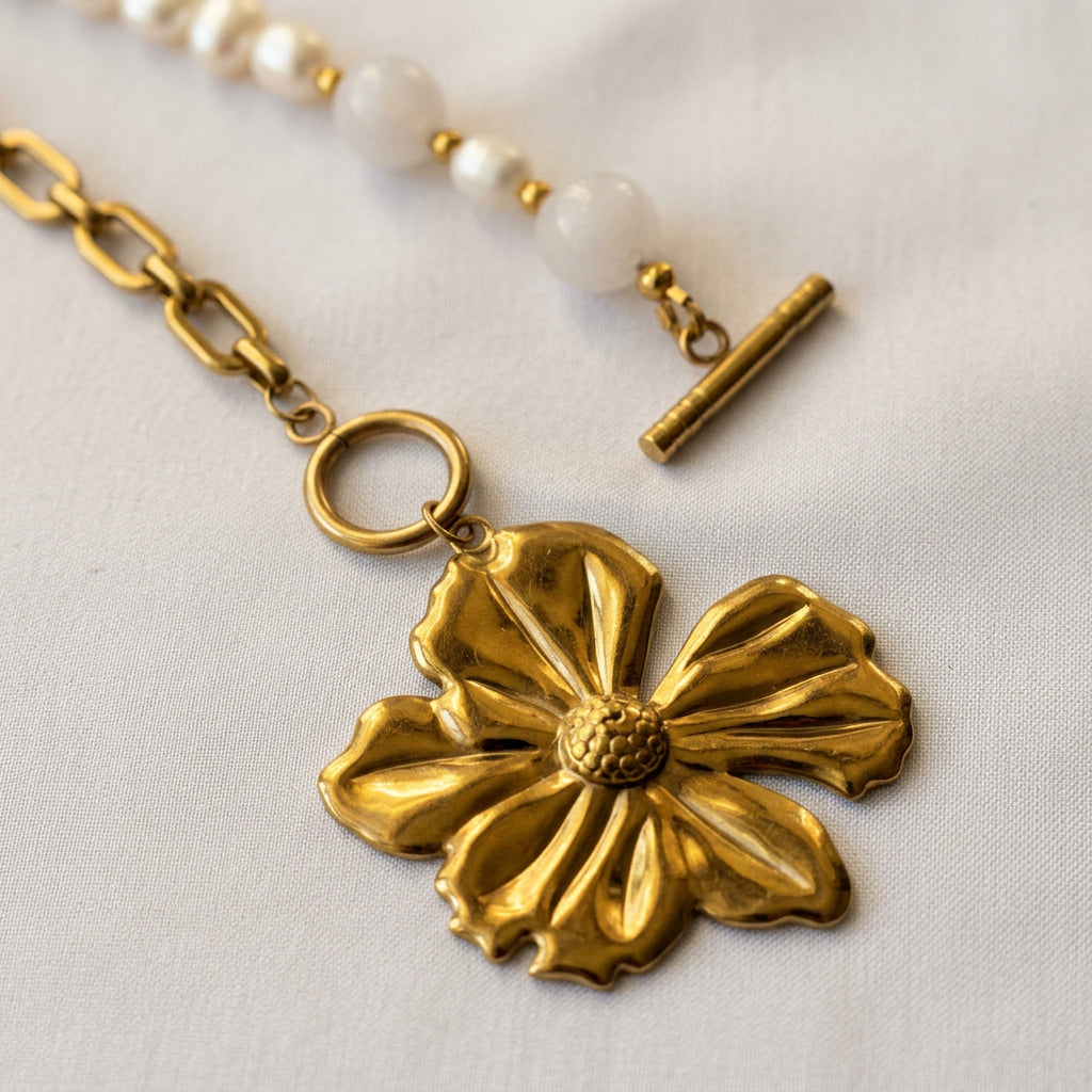 Close-up of an unfastened gold-plated toggle clasp, showing the separate bar and circular ring above a five-petal flower pendant, with pink quartz, white baroque pearls and paperclip chain on cream fabric.