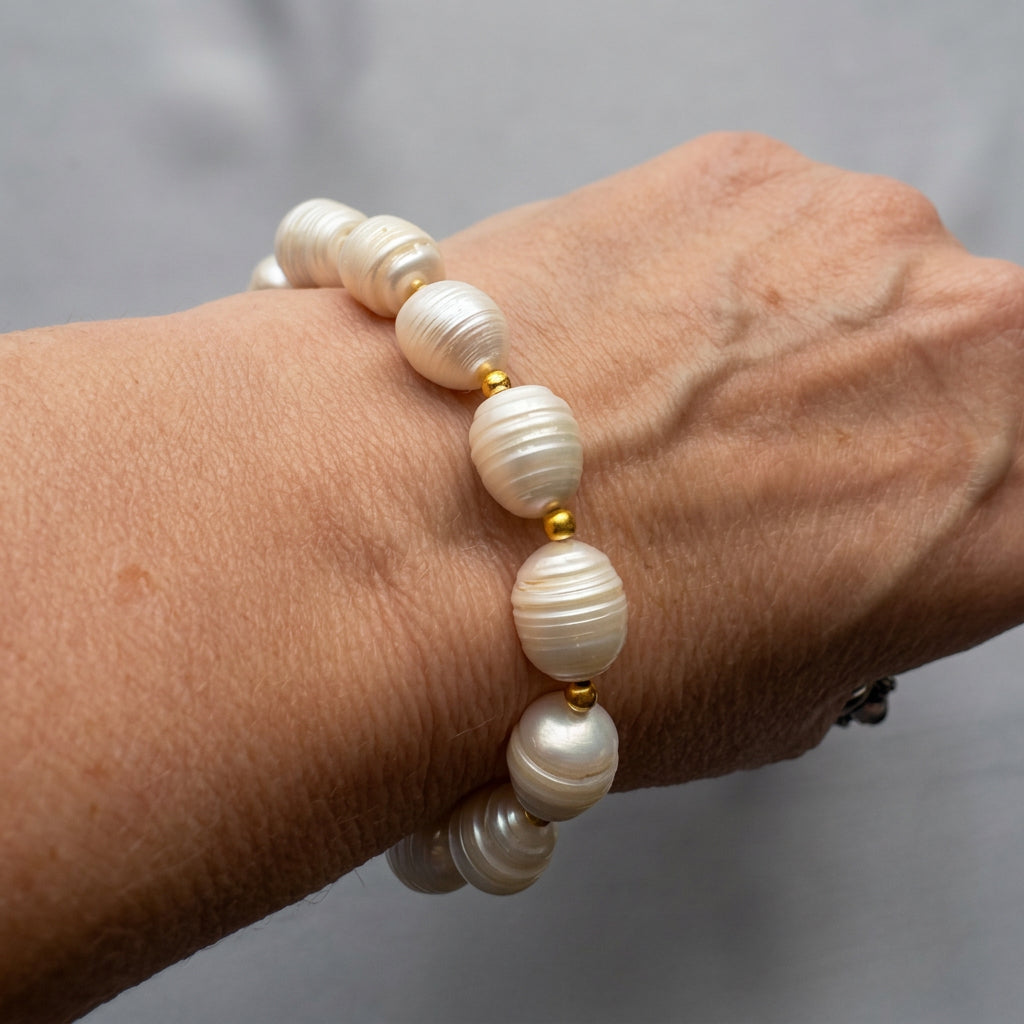 White baroque pearl bracelet worn on a wrist, showing large oval pearls with natural ridges and soft lustre, separated by small gold-coloured beads.
