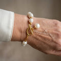 White baroque pearl bracelet with gold-coloured spacers, a fastened oval toggle clasp and a dangling white pearl charm, worn on the wrist beside a white silk shirt cuff.